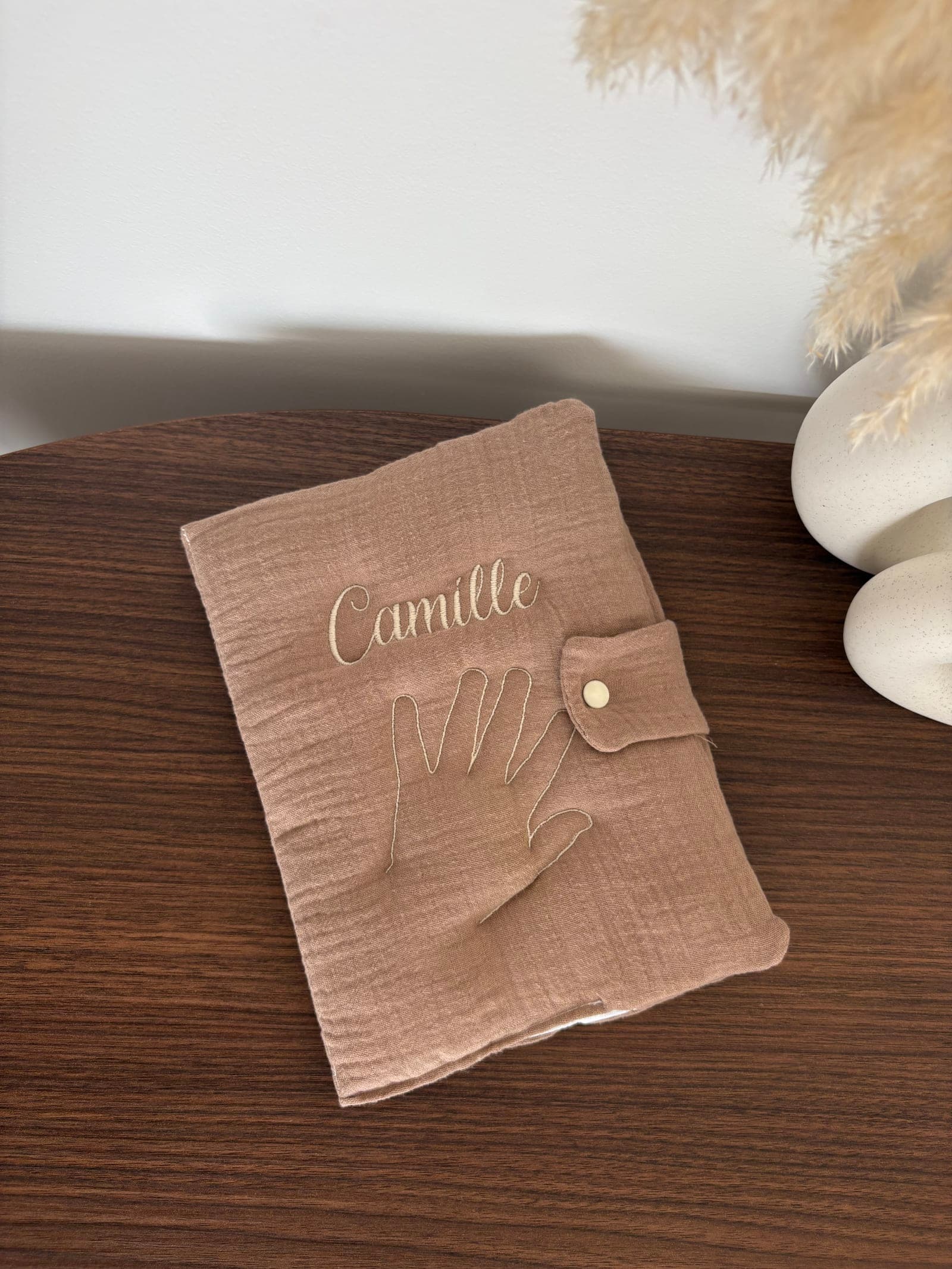 Un protège-carnet de santé en double gaze rose, brodé du prénom Camille et de l'empreinte d'une main d'enfant au fil crème.